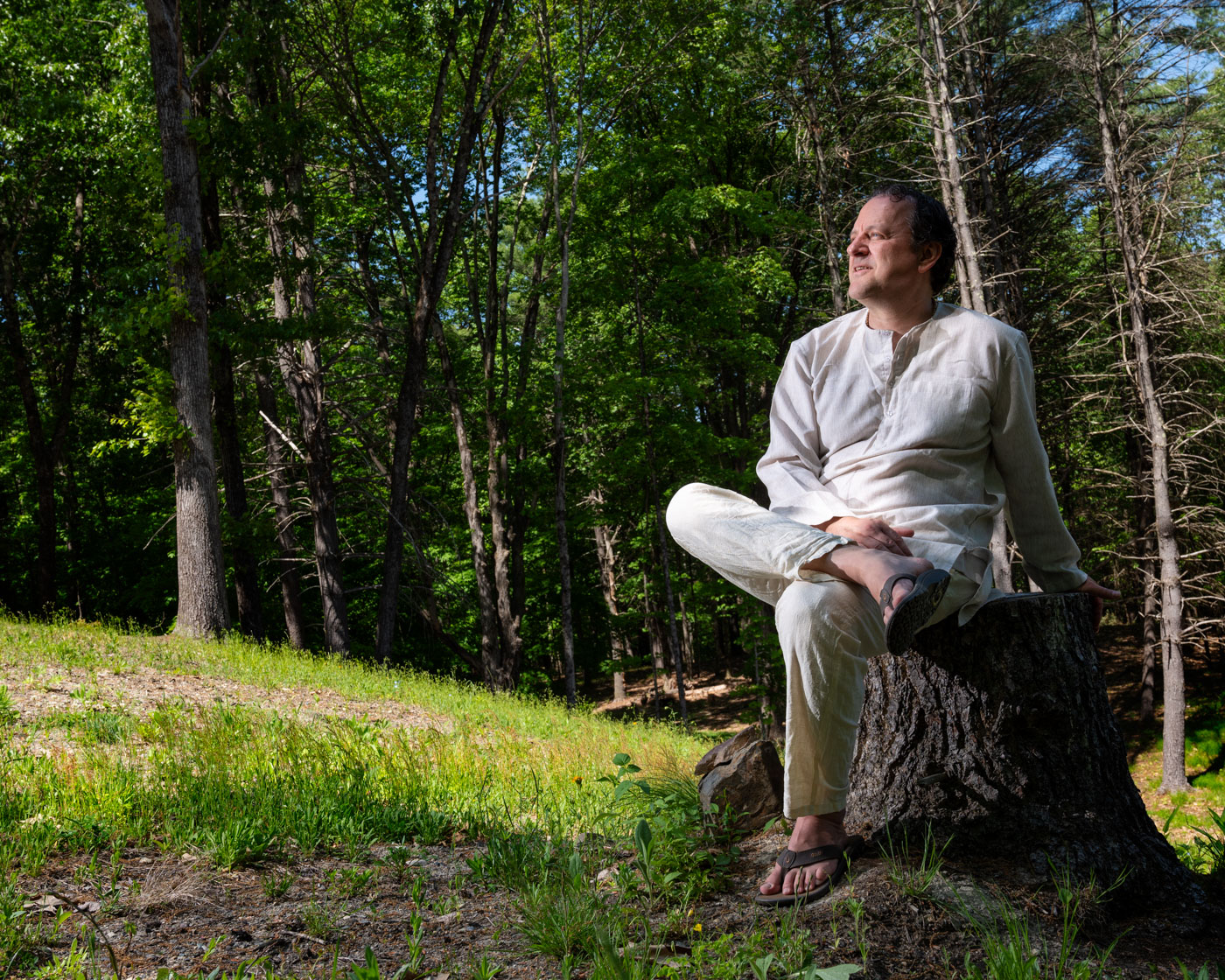 Adam Bauer in a white kurta, seated on a tree stump in a sunlit forest